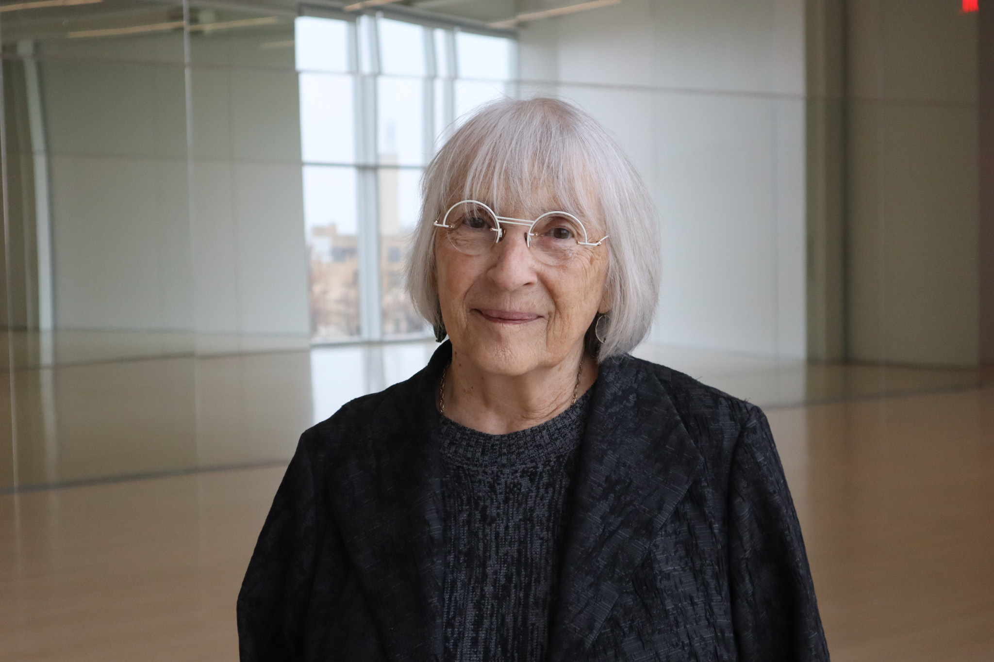 An older white woman with short white hair, white circular glasses, dressed in dark gray smiles softly, standing in a room with mirrors.