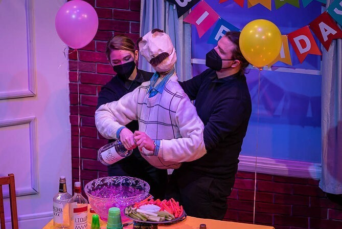 Two people dressed in black are operating a large puppet that is dressed in a striped shirt. The puppet is holding a bottle of liquor, appearing to pour into a punch bowl. Behind them are colorful balloons and a colorful sign that says birthday.