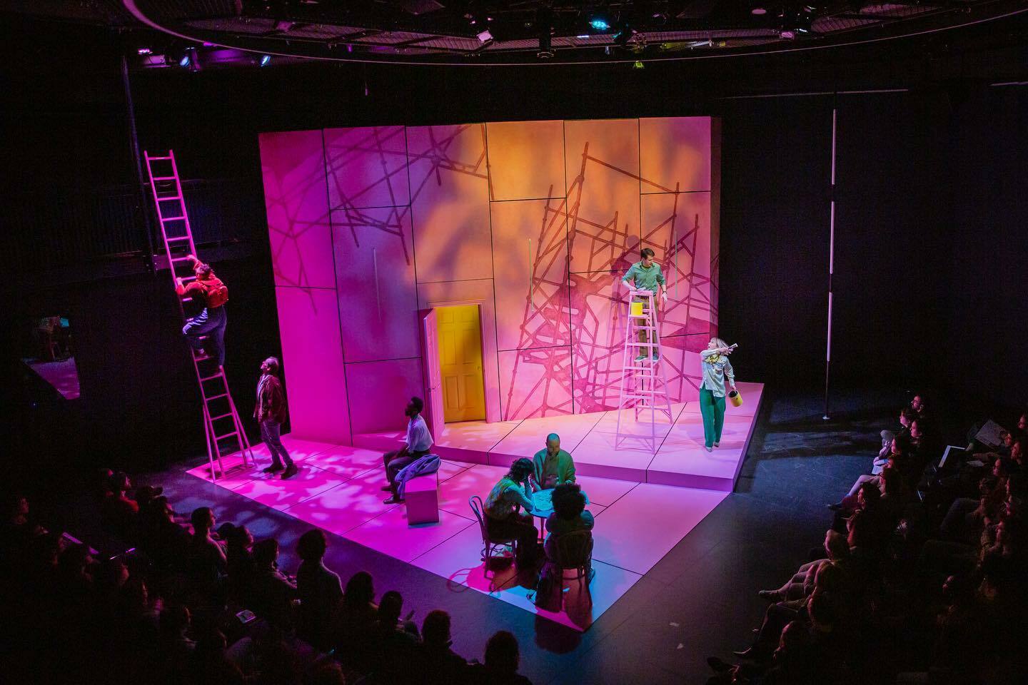 A pink set sits in the middle of an otherwise black and shadowed theater. A ladder leans against railing to the left and a door is in the center of the stage. Several actors are scattered around, standing on the pink floor.