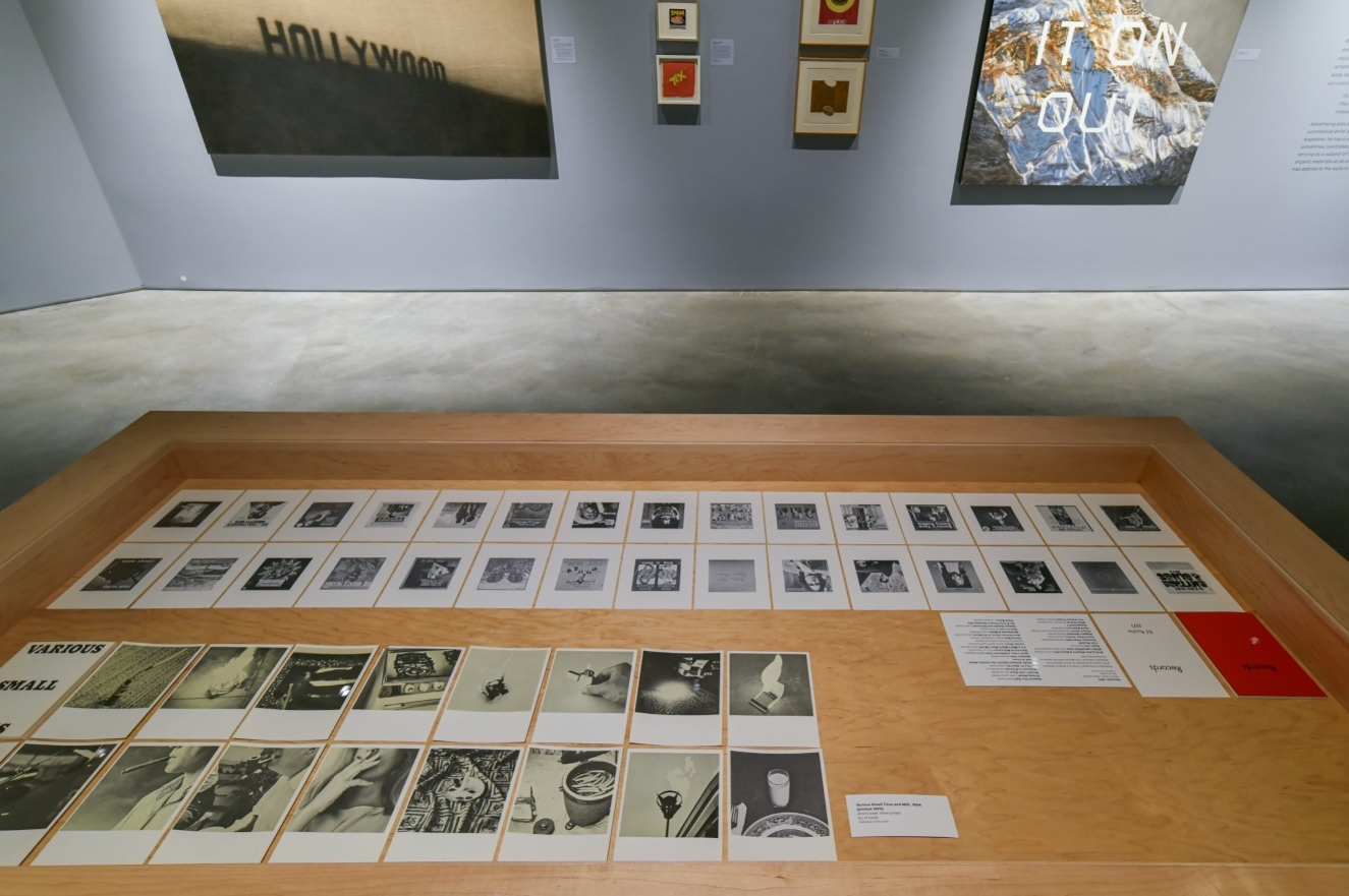 Installation view of an art gallery exhibition featuring individual printed sheets of paper on display inside a table in the foreground with larger paintings in the background