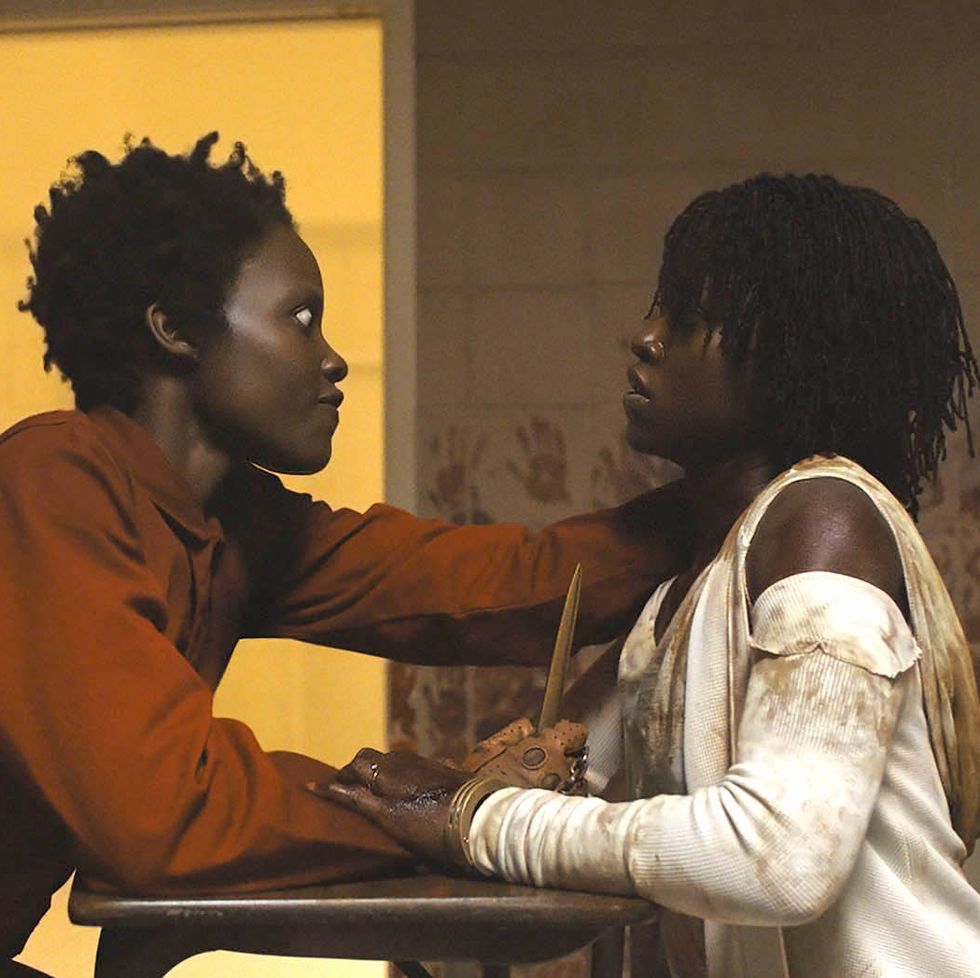 A Black person with short hair in an orange jumpsuit holds another Black figure, dressed in white, by the neck
