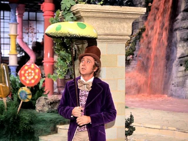 A man dressed in a purple velvet suit jacket with a brown top hat is standing in front of a chocolate waterfall and lollipop flowers. He is holding a large green mushroom cap that's stuck on the end of a pole.