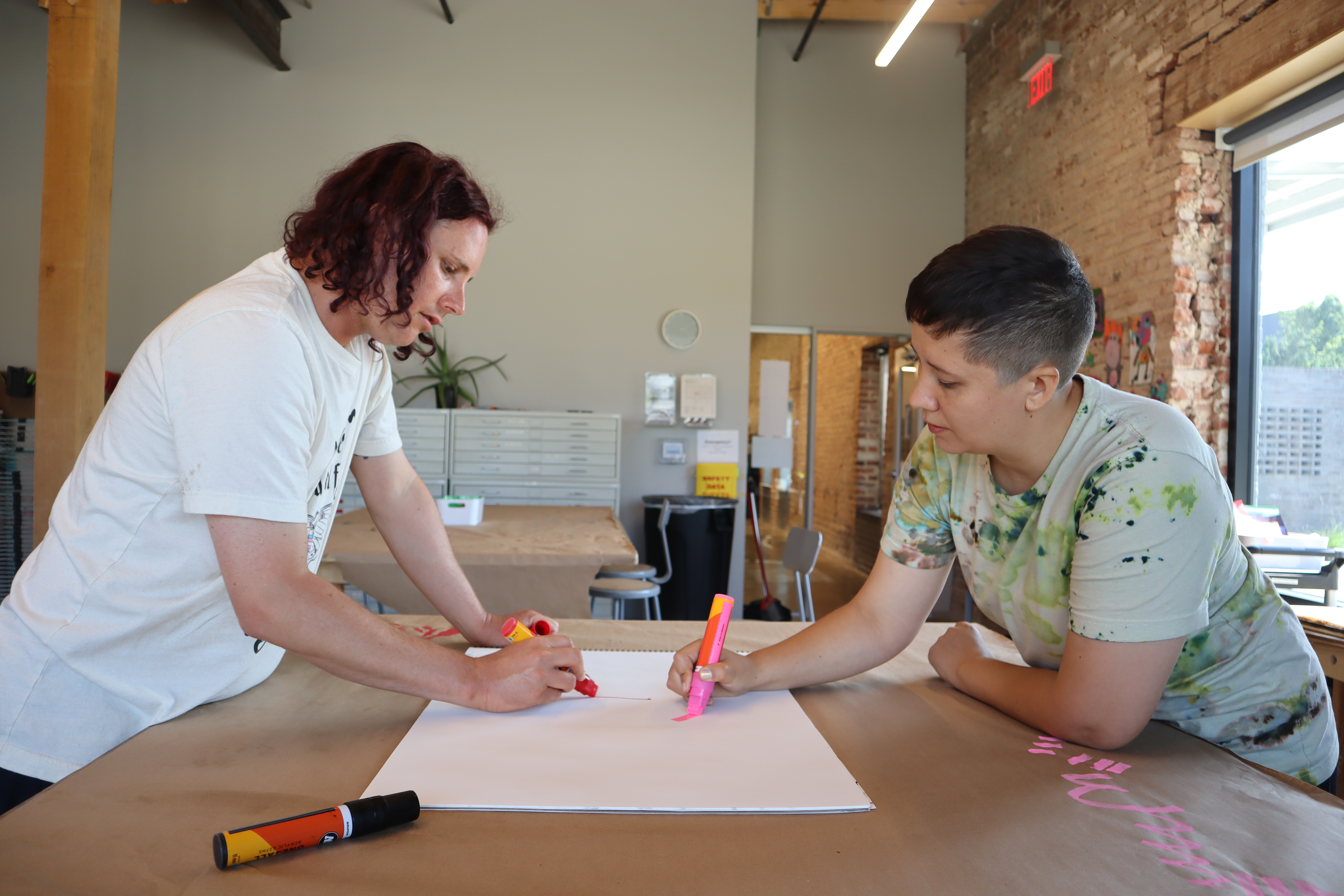 Two people stand on opposite sides of a large table covered in craft paper. They are leaning over, each holding a different colored marker, and are drawing on a large white piece of paper between them.