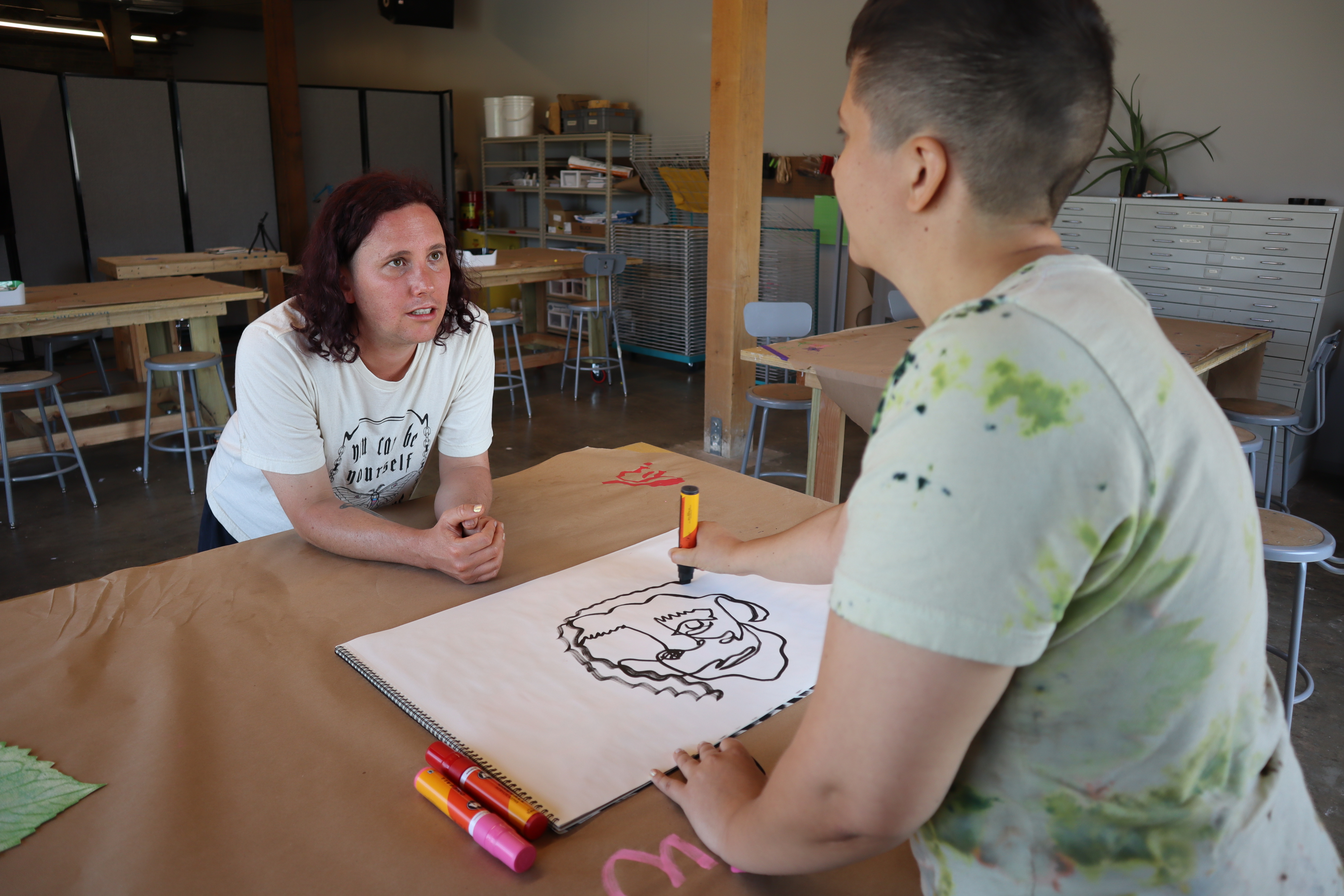 Two people stand on opposite sides of a table. We are looking over the shoulder of one as they draw a portrait of the person across from them in black marker on a large white piece of paper. The person being drawn is looking up.
