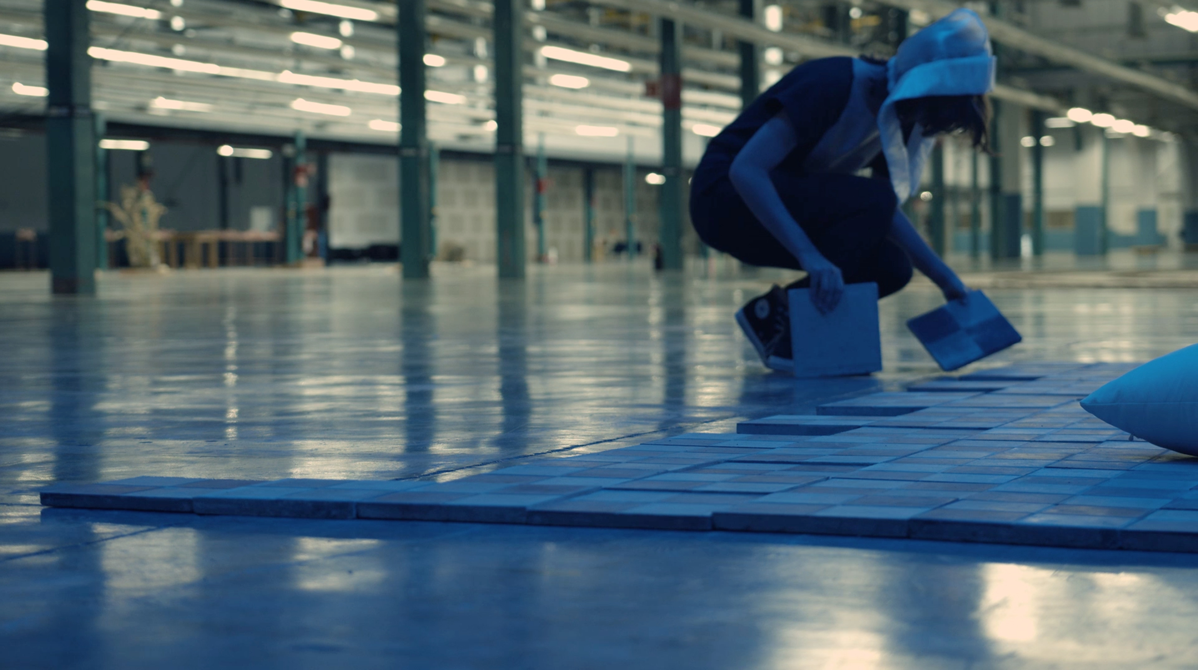 A person with a white head covering is leaned over tiles on the floor. Blue light floods in. The background looks like a warehouse with tall pillars.