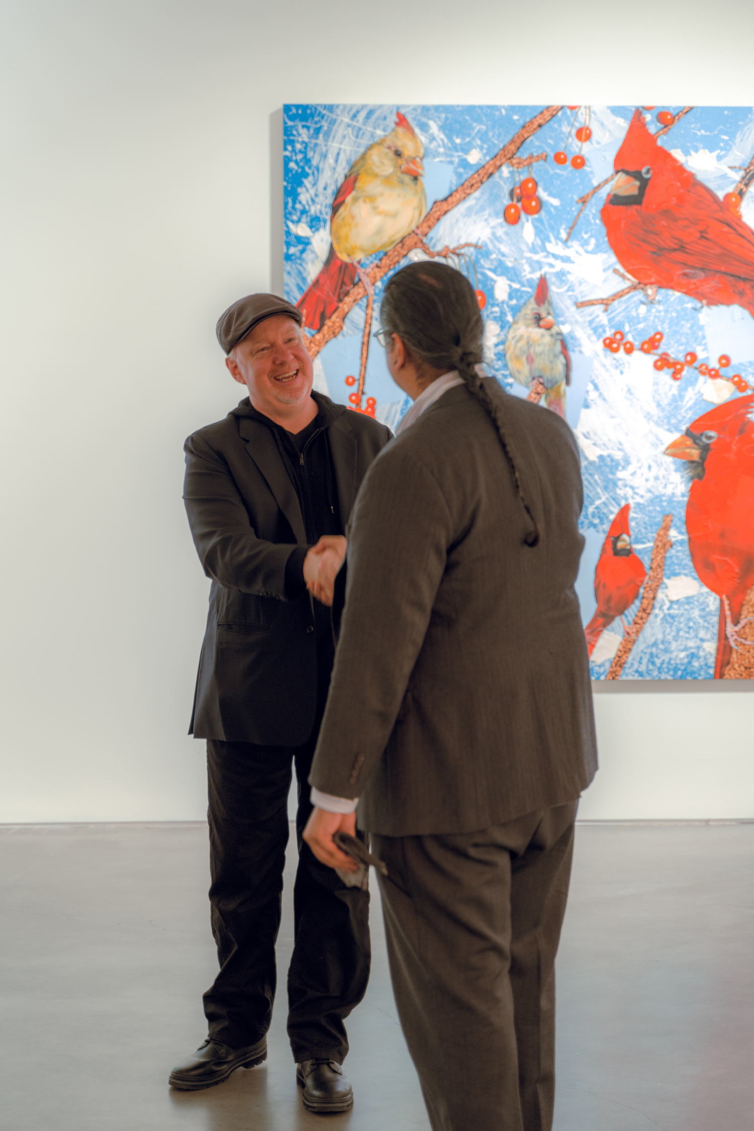 Two men are shaking hands, one facing us with a big smile and a black beret and jacket. The other is facing away from the camera, in a tweed suit and braided hair. In the background is a large painting of red cardinals on a blue background.