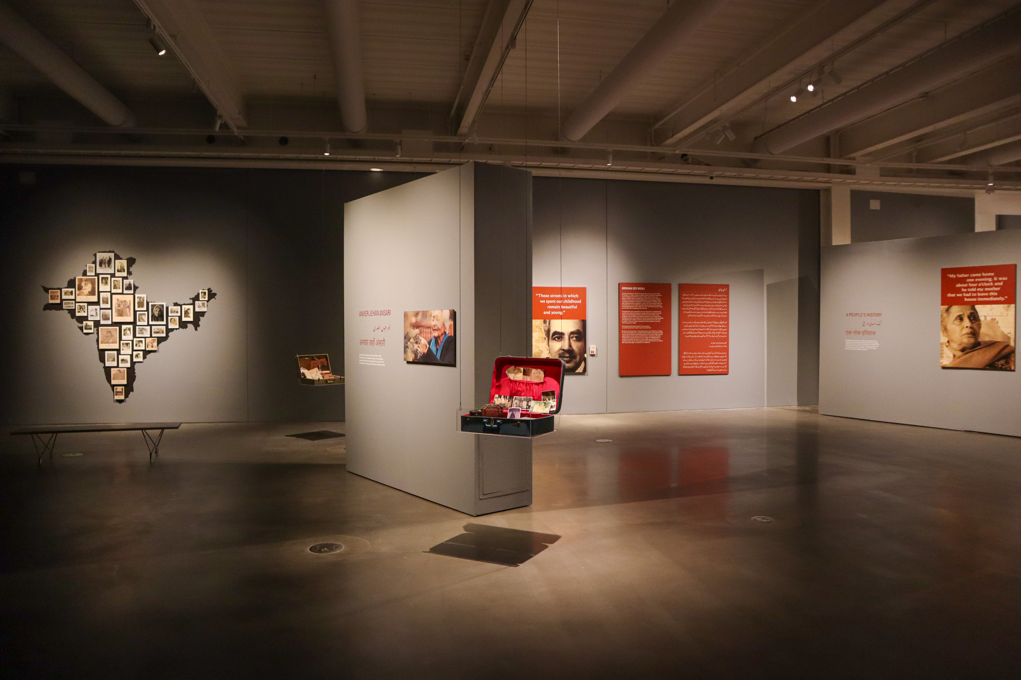 An art gallery shows a suitcase hanging from the ceiling in the middle of a room with a map of India to the left, and orange boards with white writing to the right
