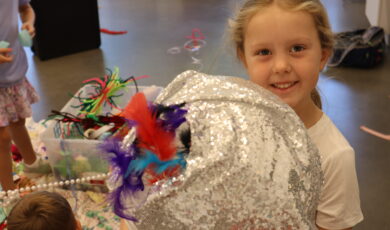 A child holds a shiny silver sphere with colorful feathers on top. Additional craft supplies are on a table in the background.