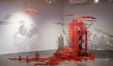 An art installation comprised of intricately cut red and orange screens spread across the floor, standing and suspended from the ceiling of a gallery