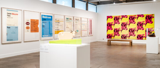 A gallery filled with food-related art, including a pink and yellow cow painting, a sculpted cow on a plinth and prints depicting food as medicine labels
