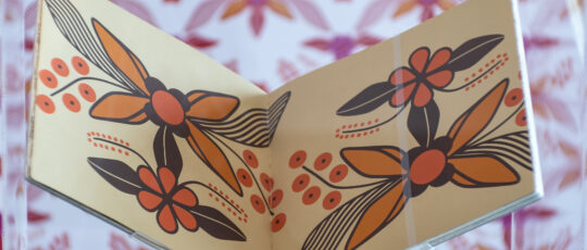 A book with a large floral pattern is held open on a stand in front of similar wallpaper