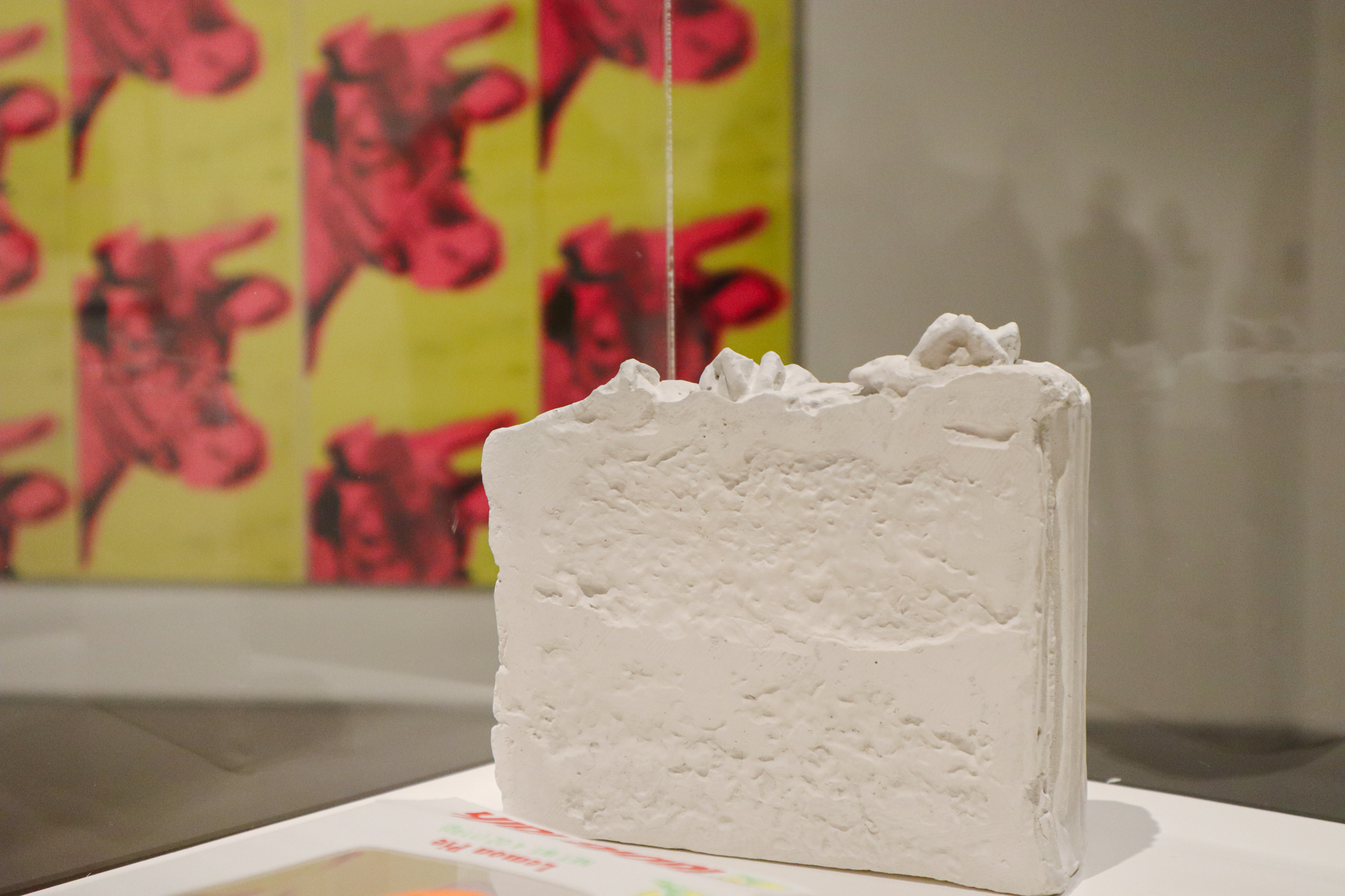 A ceramic slice of white cake sits on a white plinth with a glass case around it. We can see a large piece behind the cake with pink cow heads printed over and over on a yellow background.