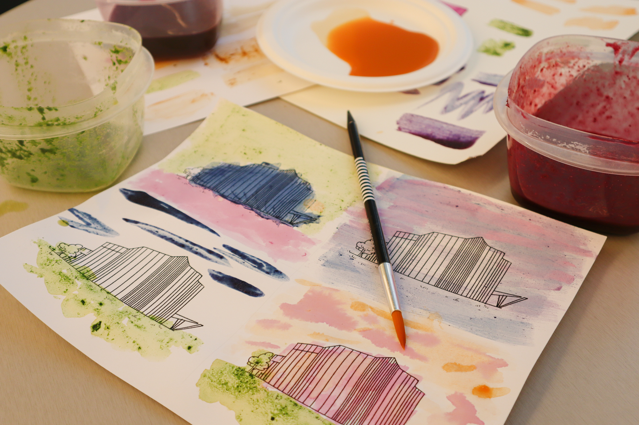 A watercolor painting of four buildings sits on a table. We can see containers of green, purple and orange ink in containers. A black paintbrush sits on the watercolor.