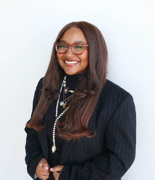 A Black woman in a black blaser and variety of low and high necklaces, with dark brown hair and red-rimmed glasses, smiles at the camera