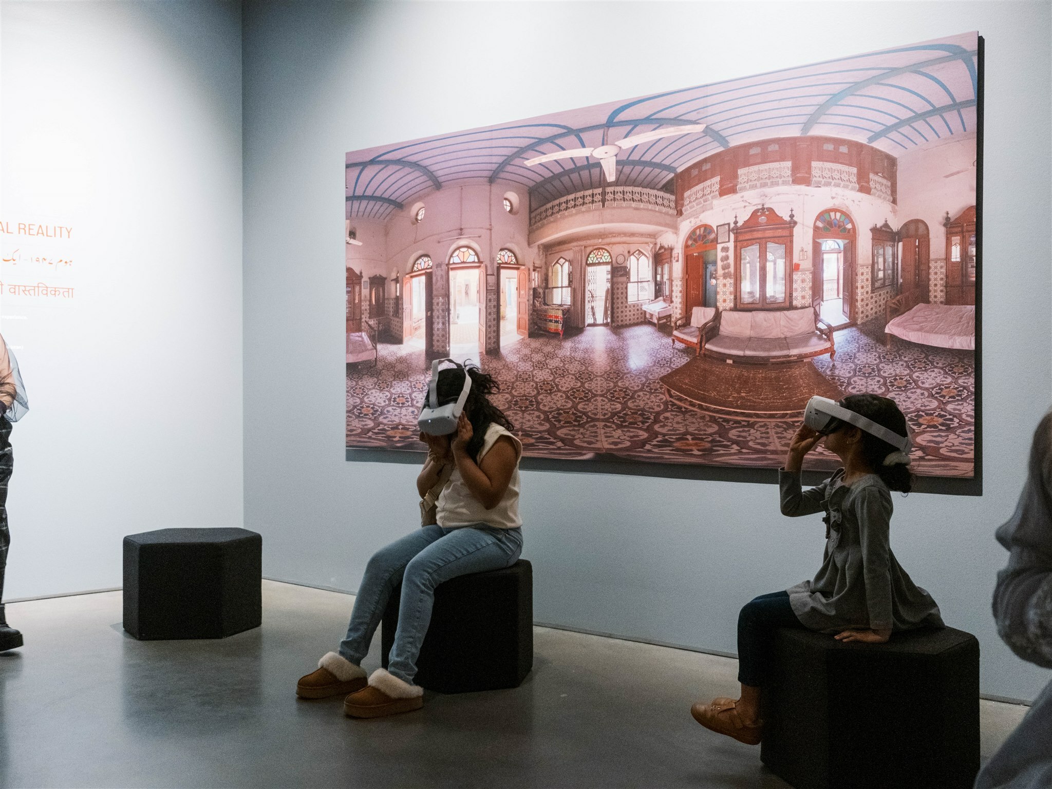 Two children sit on circular black stools in front of a large painting of a colorful interior hospital in Pakistan. The two kids have virtual reality headsets on.