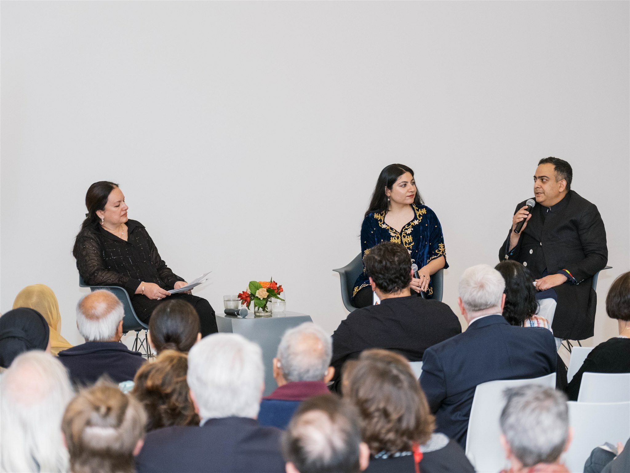 Three people sit on a stage against a white wall, all with dark hair and of South Asian decent, all dressed in black. A crowd of people sit in front of them.
