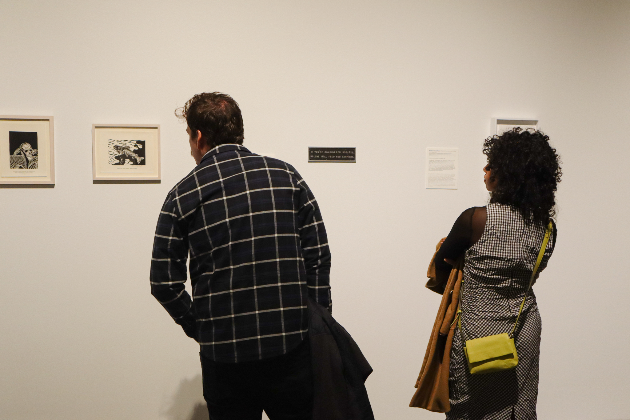 Two people are looking at art hanging on a white wall. There are three visible pieces: two black prints of a corn field with a skull and a salmon swimming, the other a black rectangle plate.