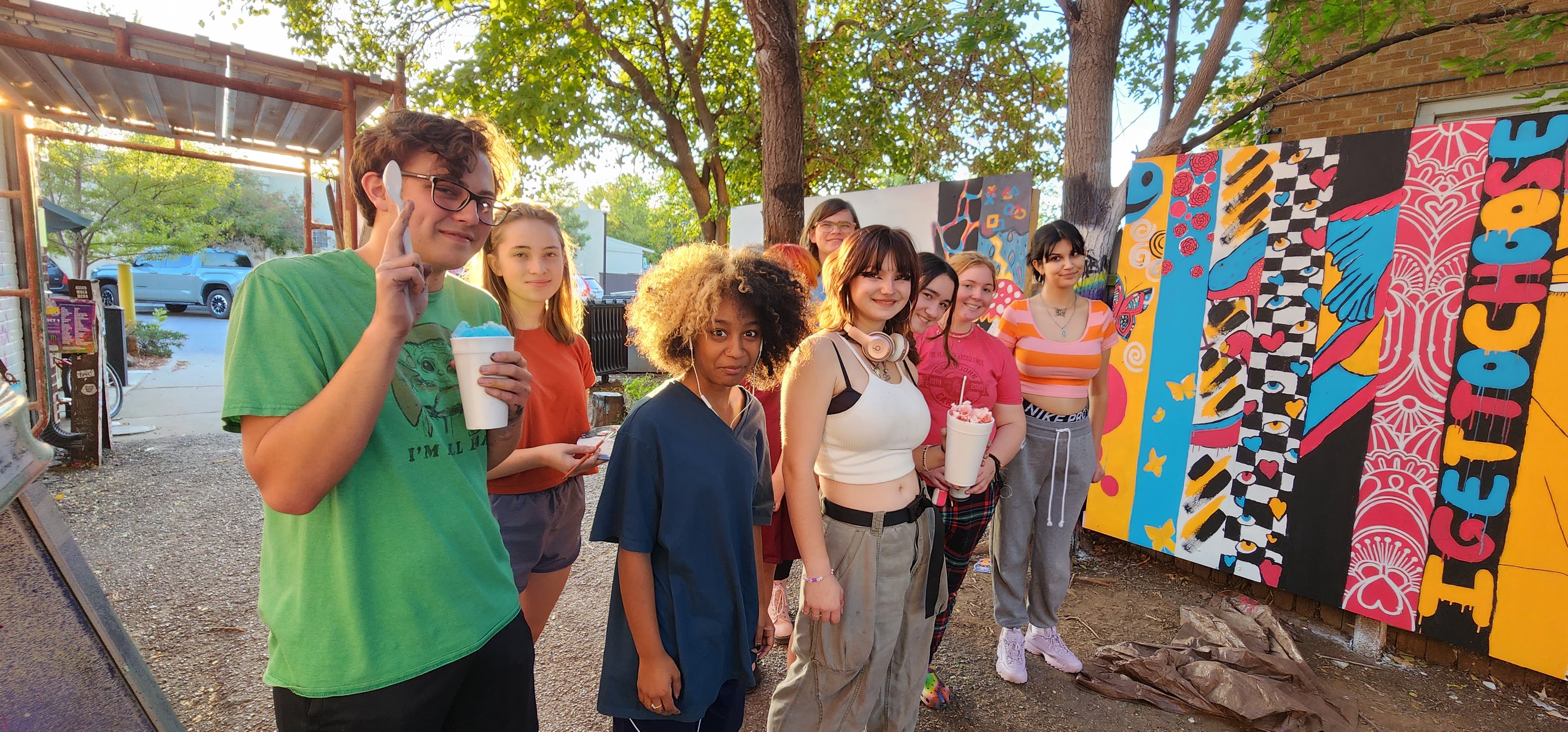 A group of teenagers stand outside next to a colorful mural. They all have snow cones and are smiling at the camera.