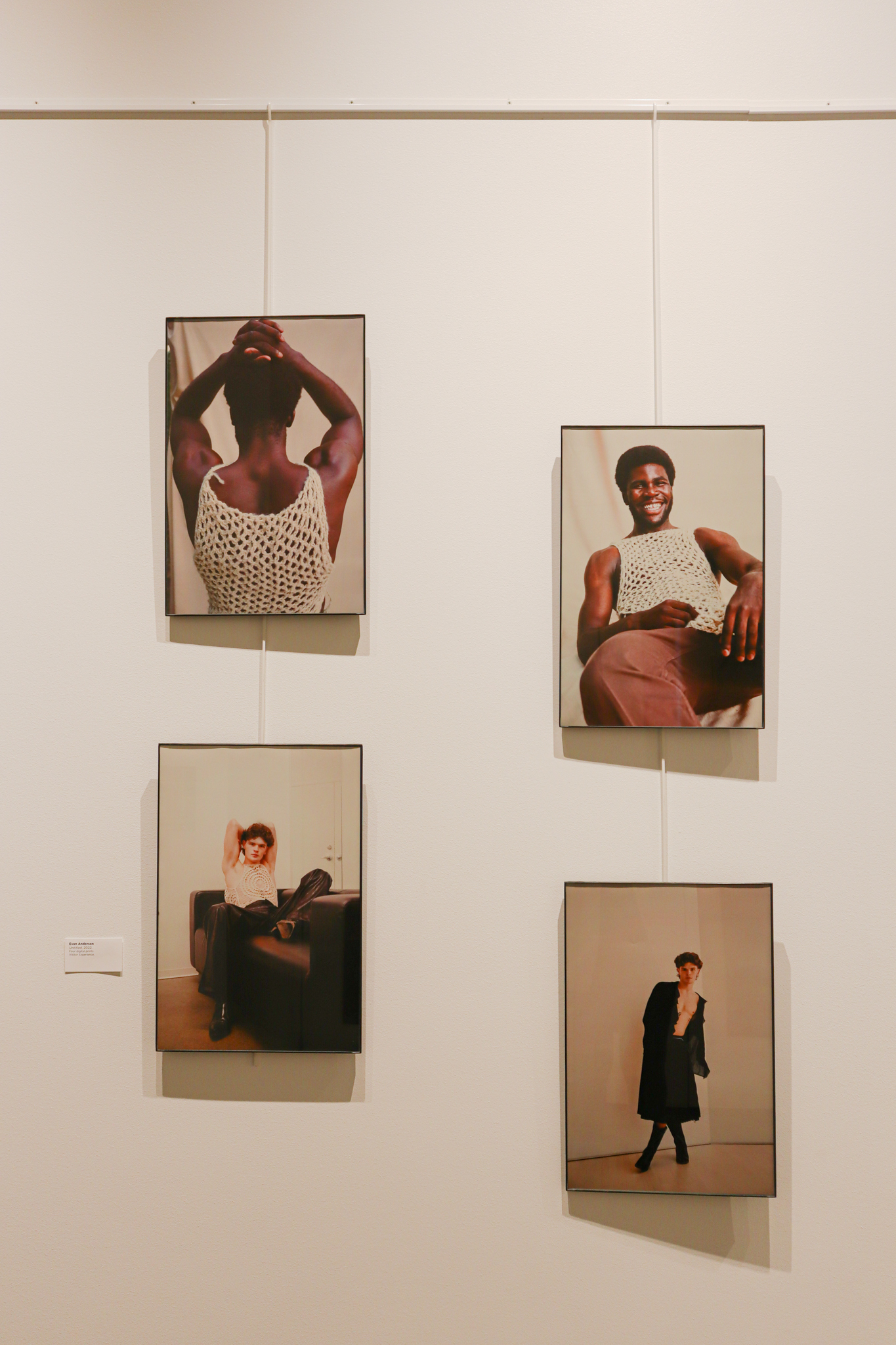 Four photographs are hung on a white wall, staggering in two rows stacked next to one another. In the top two images, a Black masculine-presenting individual is in a white crochet top. In the bottom two, a white masculine-presenting man is in a dark coat.