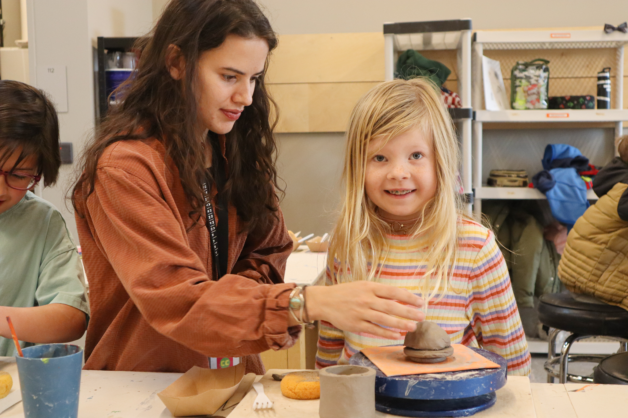 A person in a red-brown jacket with dark hair is helping a young kid with clay. The child has blonde hair is looking at the camera, smiling with braces, and is wearing a striped colorful shirt.