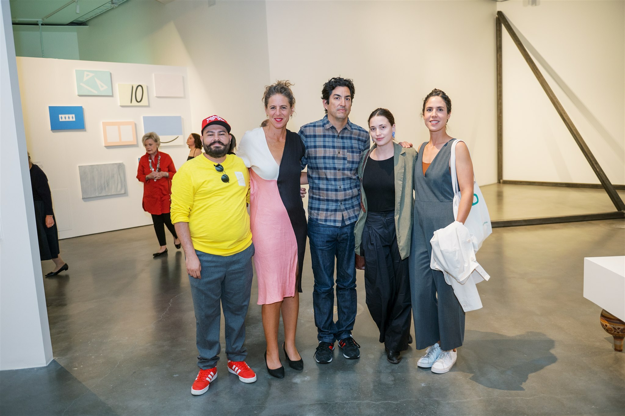 Five people stand in an art gallery looking at the camera. From left to right, there is a man in a yellow shirt, woman in a pink, black and white dress, man in a checkered button-up and two women in grey and black.