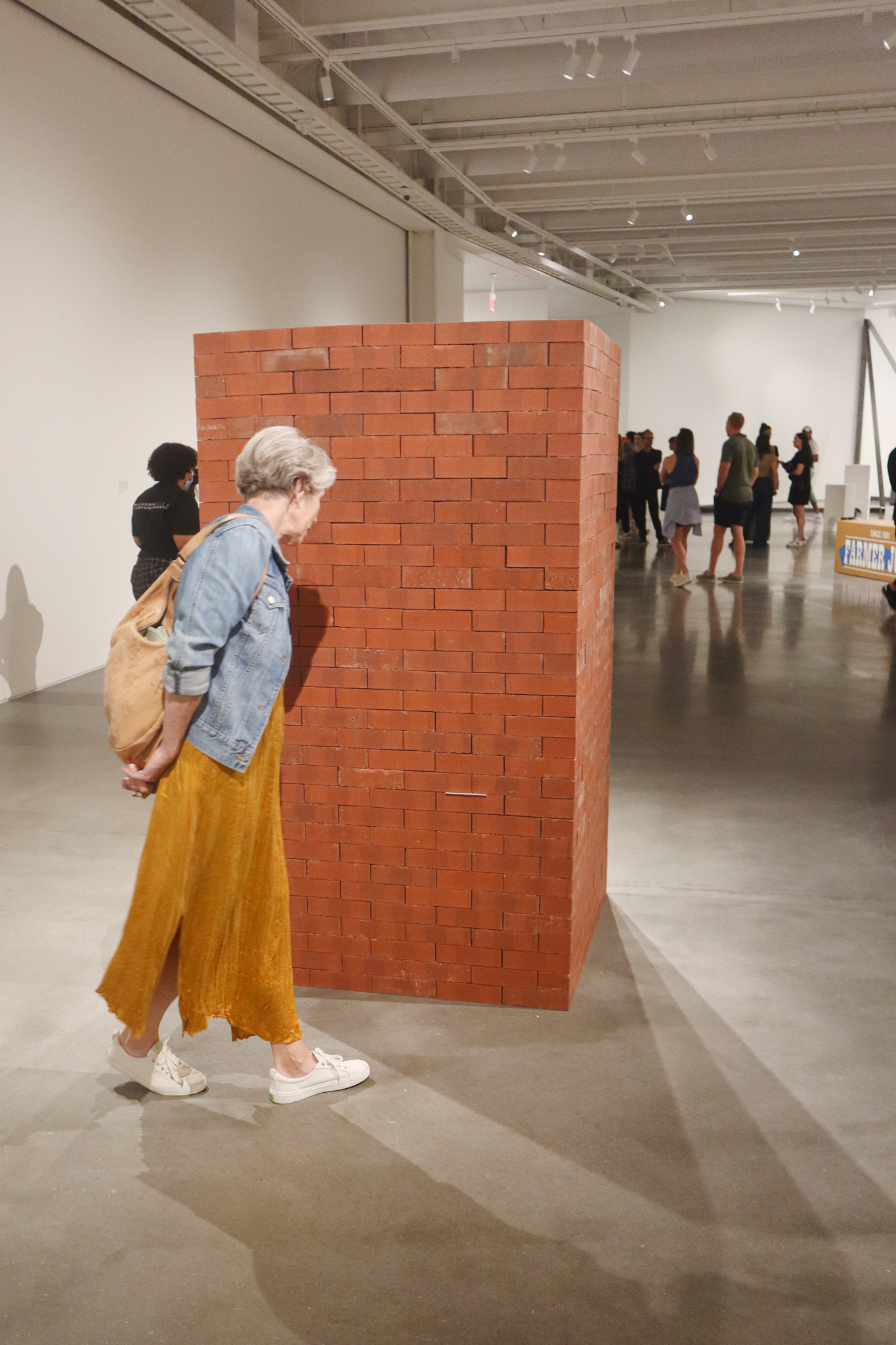 A person with a denis jacket and long, mustard-colored skirt is walking around a tall column of bricks.