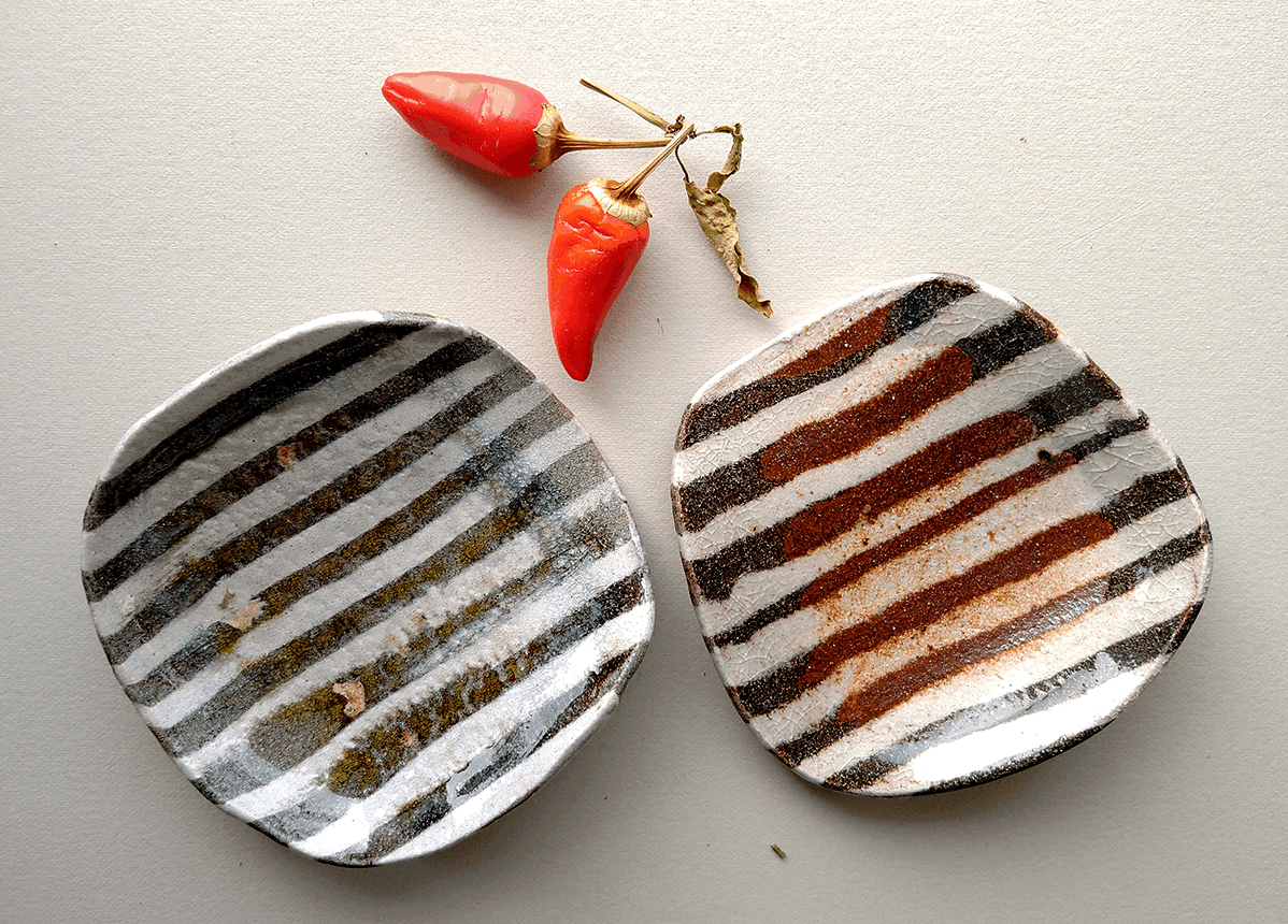 Two ceramic plates sit next to one another, with two red peppers sitting above them. The plates are a reddish-brown and white stripped pattern.