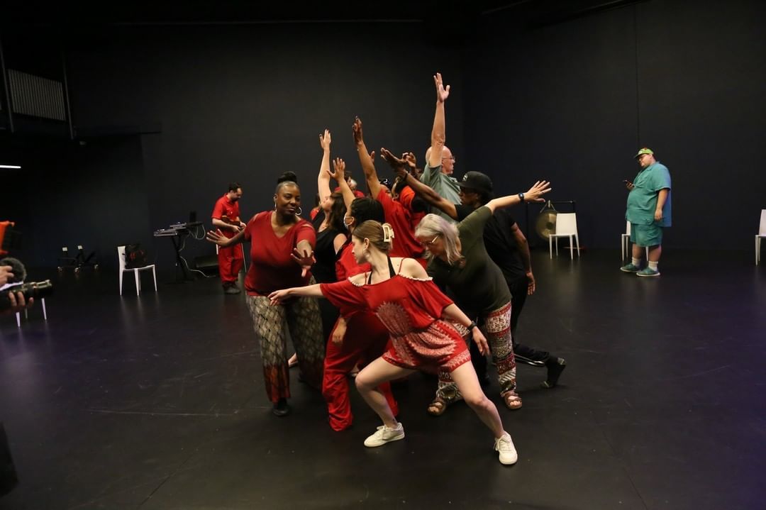 A group of dancers in a dark room are all leaning into one another, their arms stretched in all directions, forming a blob of moving dancers. All are dressed in red.
