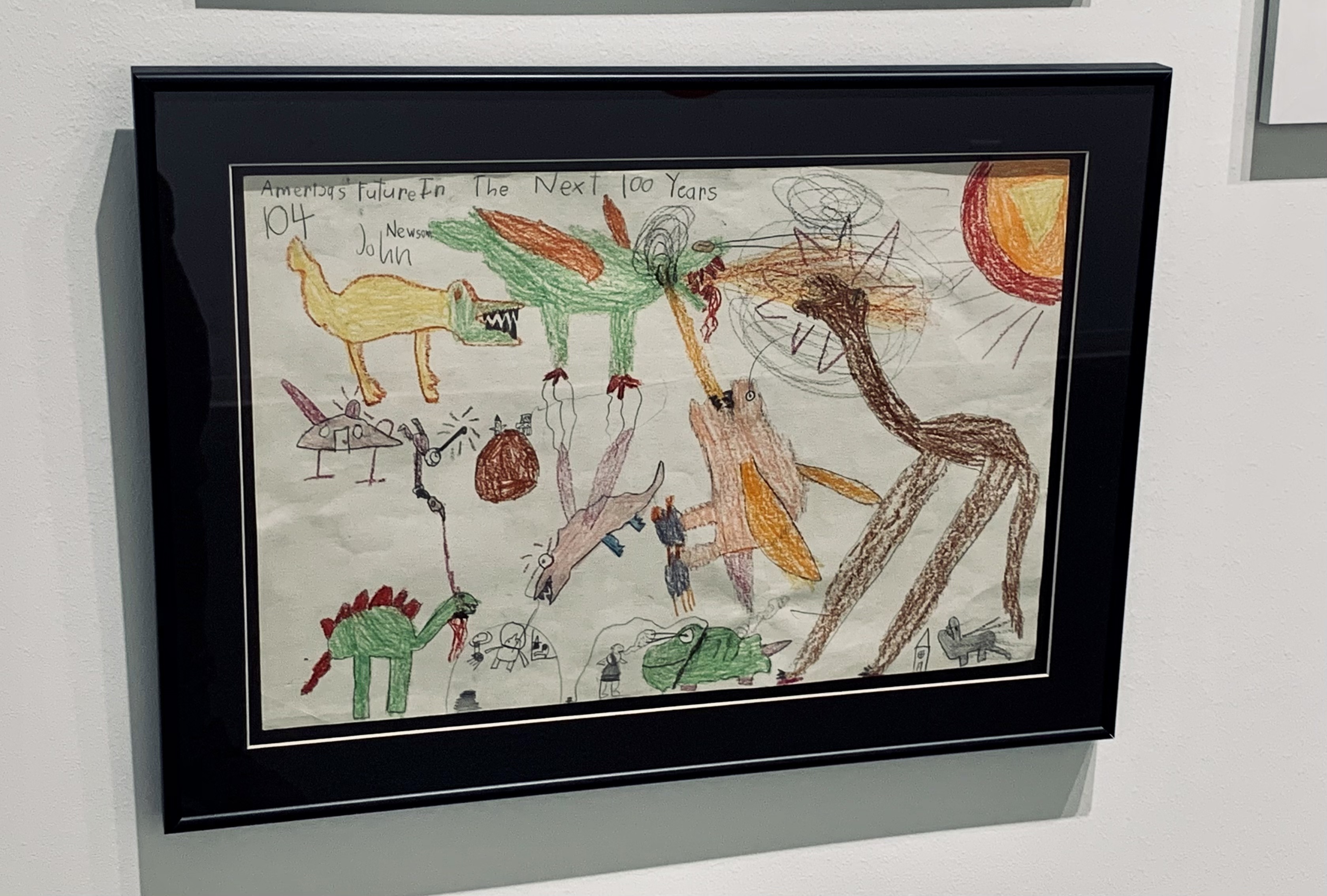A drawing in a black frame hangs on the wall. Kid-like characters are drawn, some look like dinosaurs, some flying beasts, in bright purples and reds and greens.