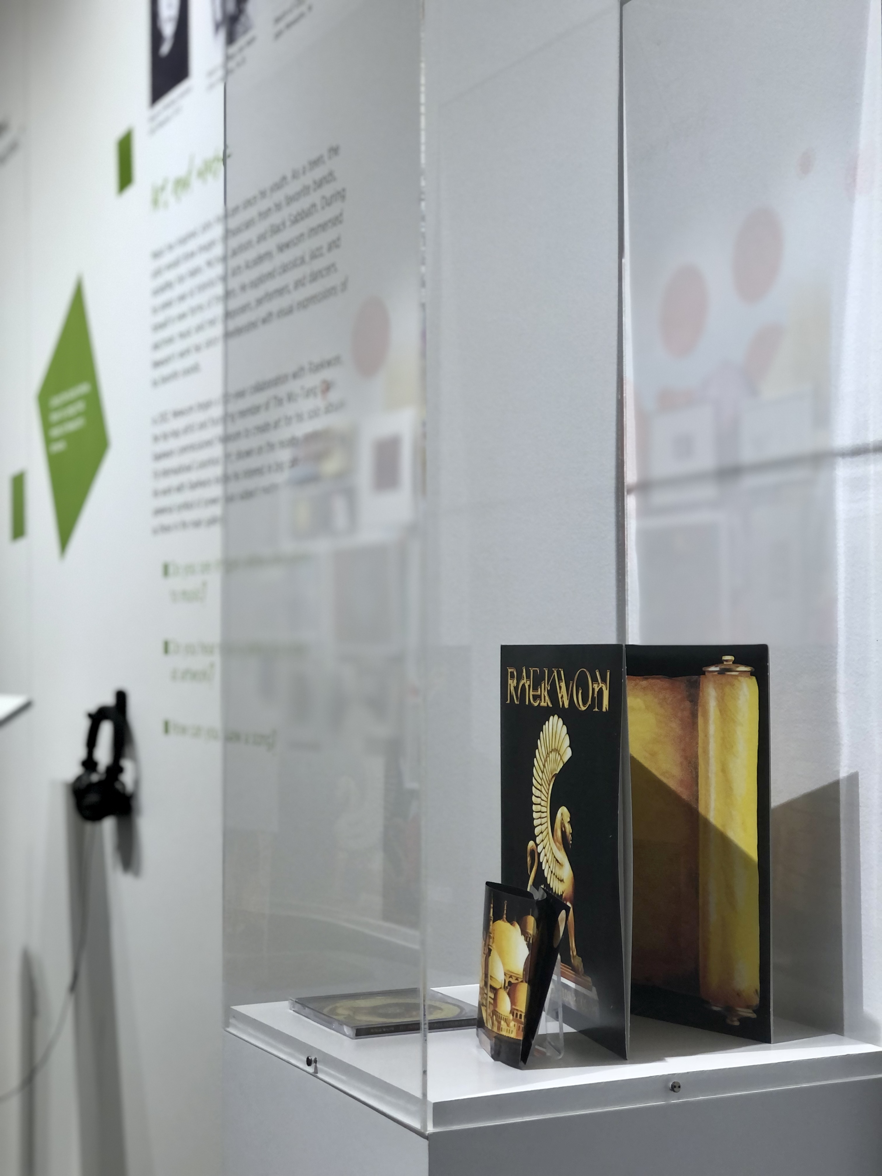 A glass case sits against a white wall. Inside is a vinyl album and CD with a black background and a golden winged creature. We can see green squares and text on the wall to the right, as well as a kiosk with headphones.