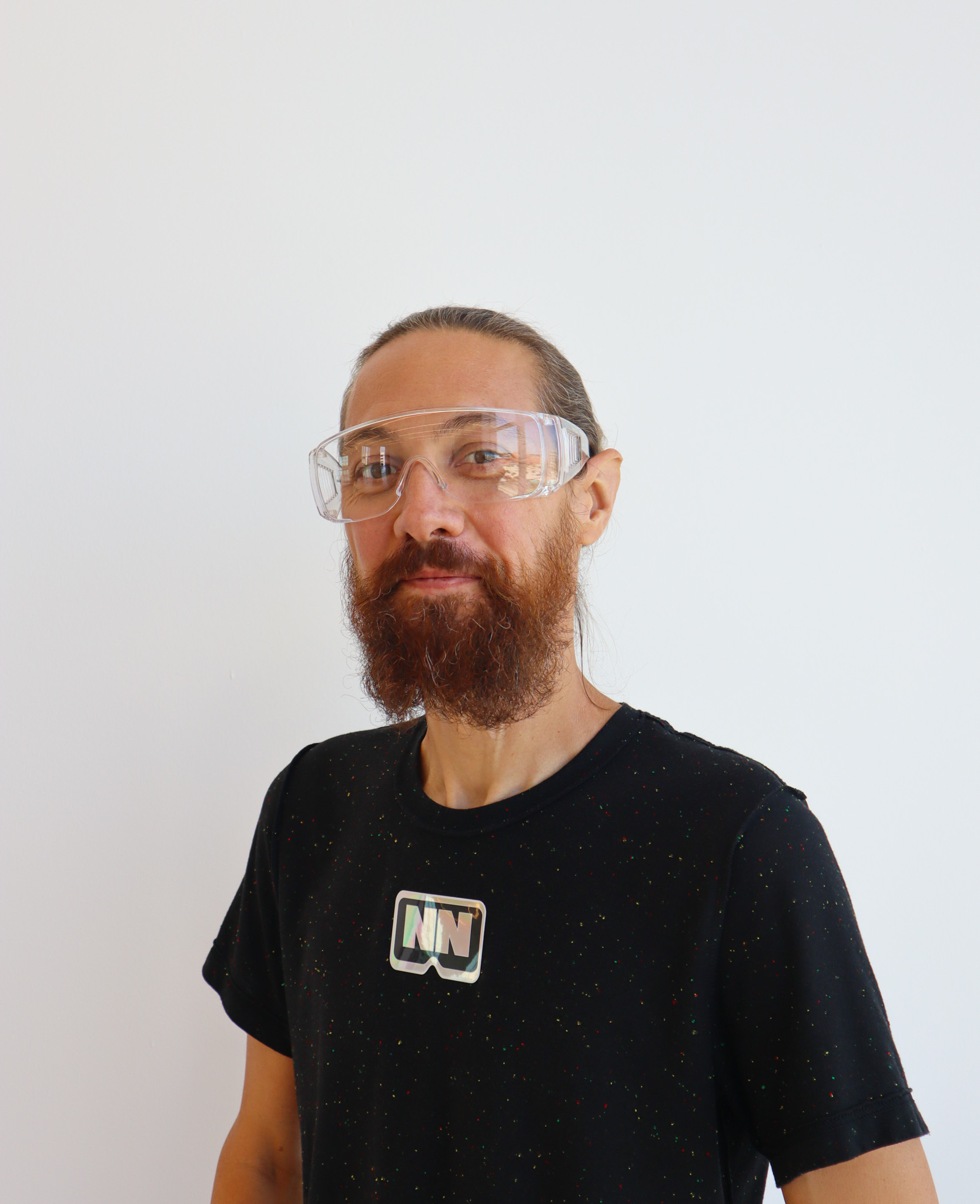 A person smiles softly at the camera, standing against a white wall. He has on large clear glasses and is wearing a dark shirt. His brown hair is pulled back, and he has a thick brown beard.