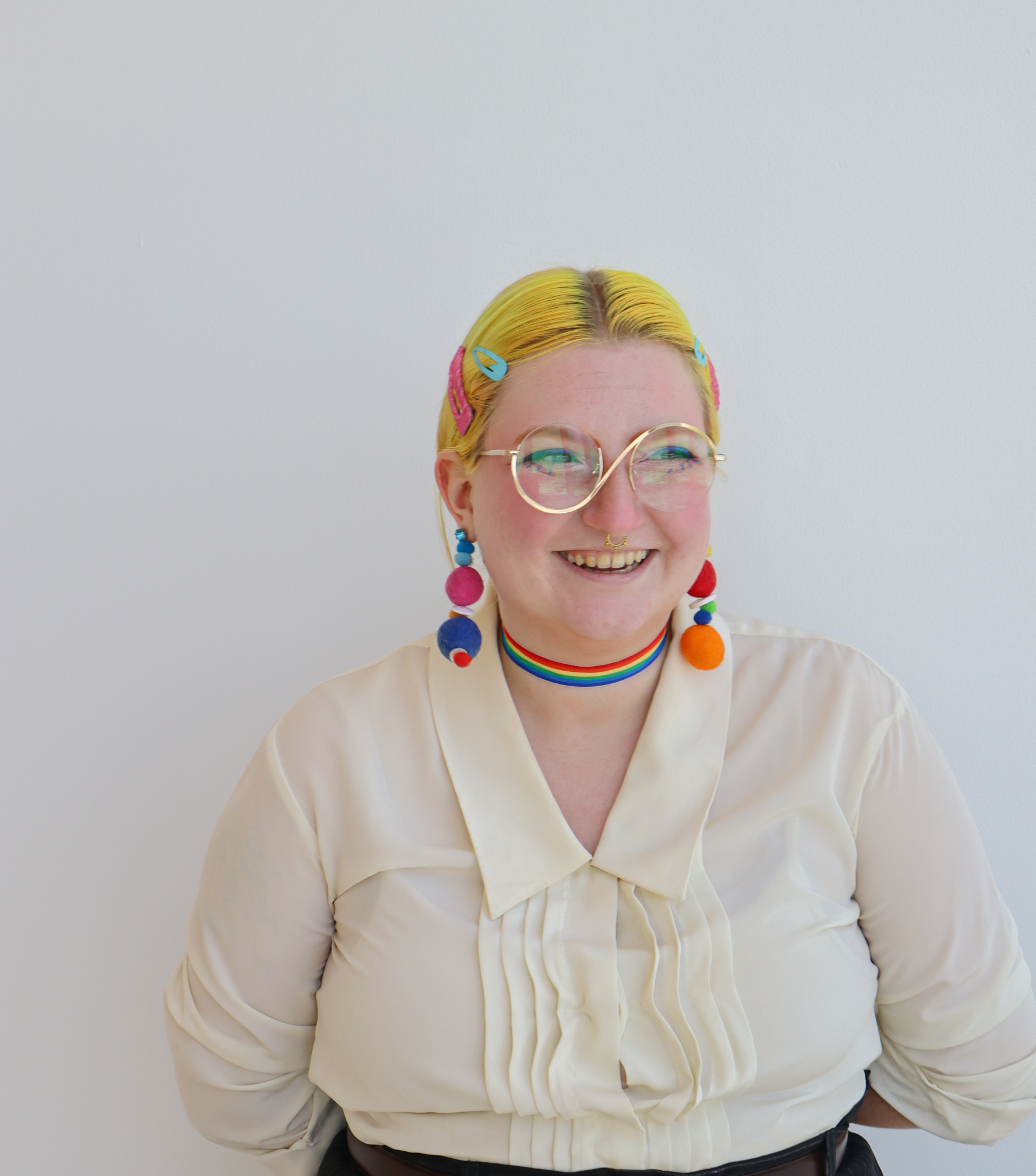 A white person with bright yellow hair and colorful makeup smiles, looking to the side They are dressed in an off-white blouse, with a rainbow choker necklace and colorful pom-pom earrings.