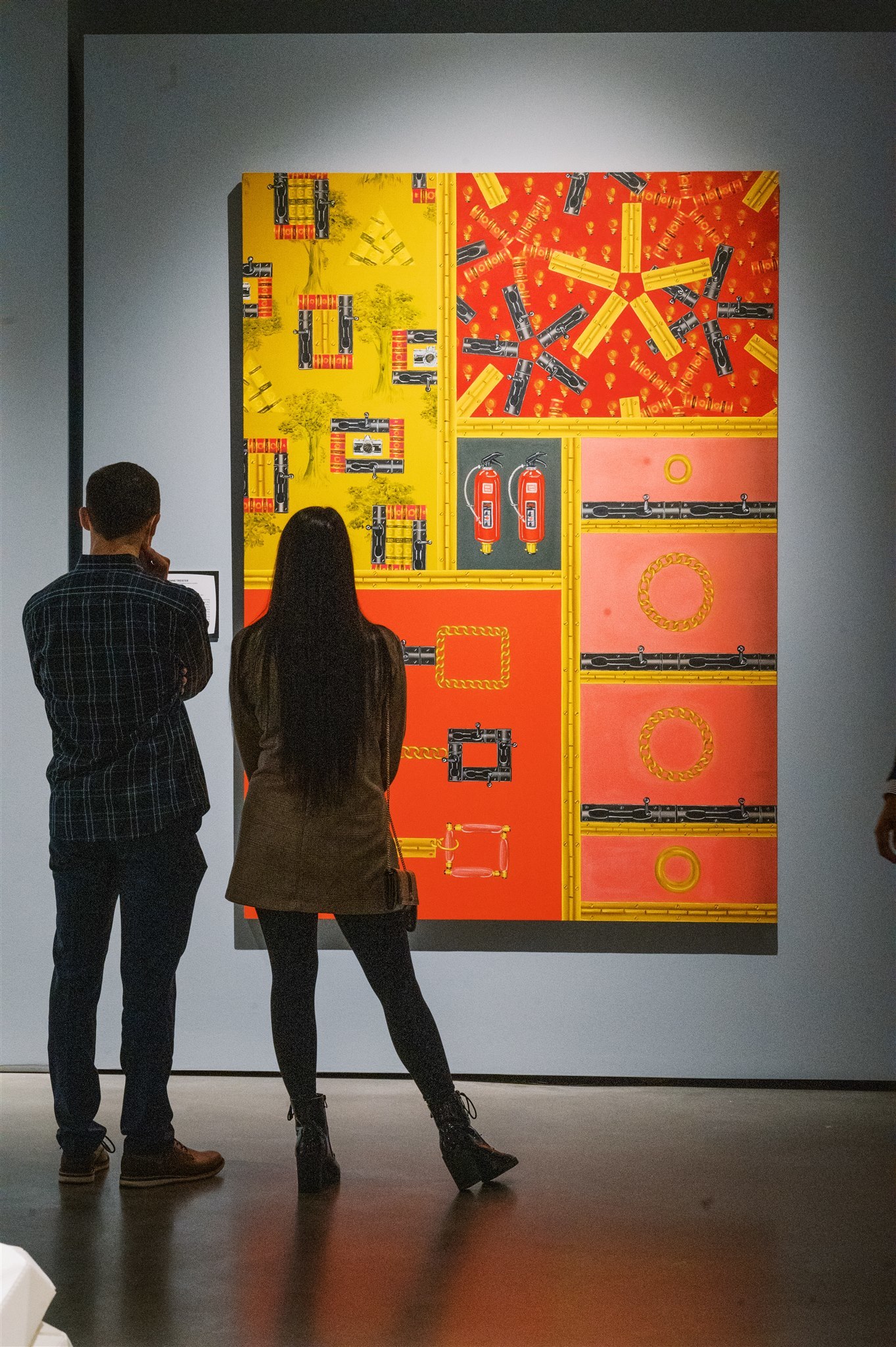 Two figures stand in a gray-walled gallery with concrete floors viewing a large painting featuring hinges, locks, trees, chains and fire extinguishers in warm yellows, reds and pinks and arranged in a grid