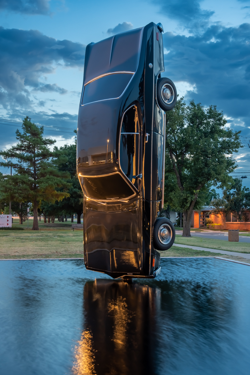 A vintage black car suspended over a reflecting pool with trees in the background