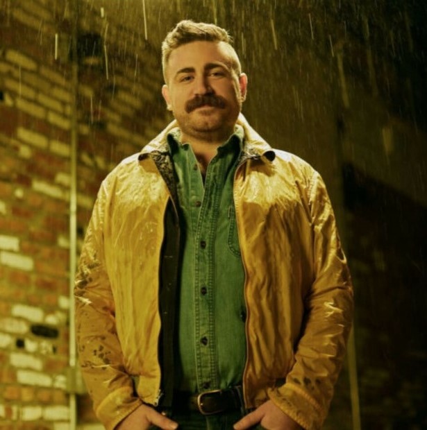 A man with a mustache wearing a yellow jacket stands in front of a brick wall