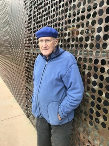An older man in a blue coat and beret stand against a wall covered in small circles