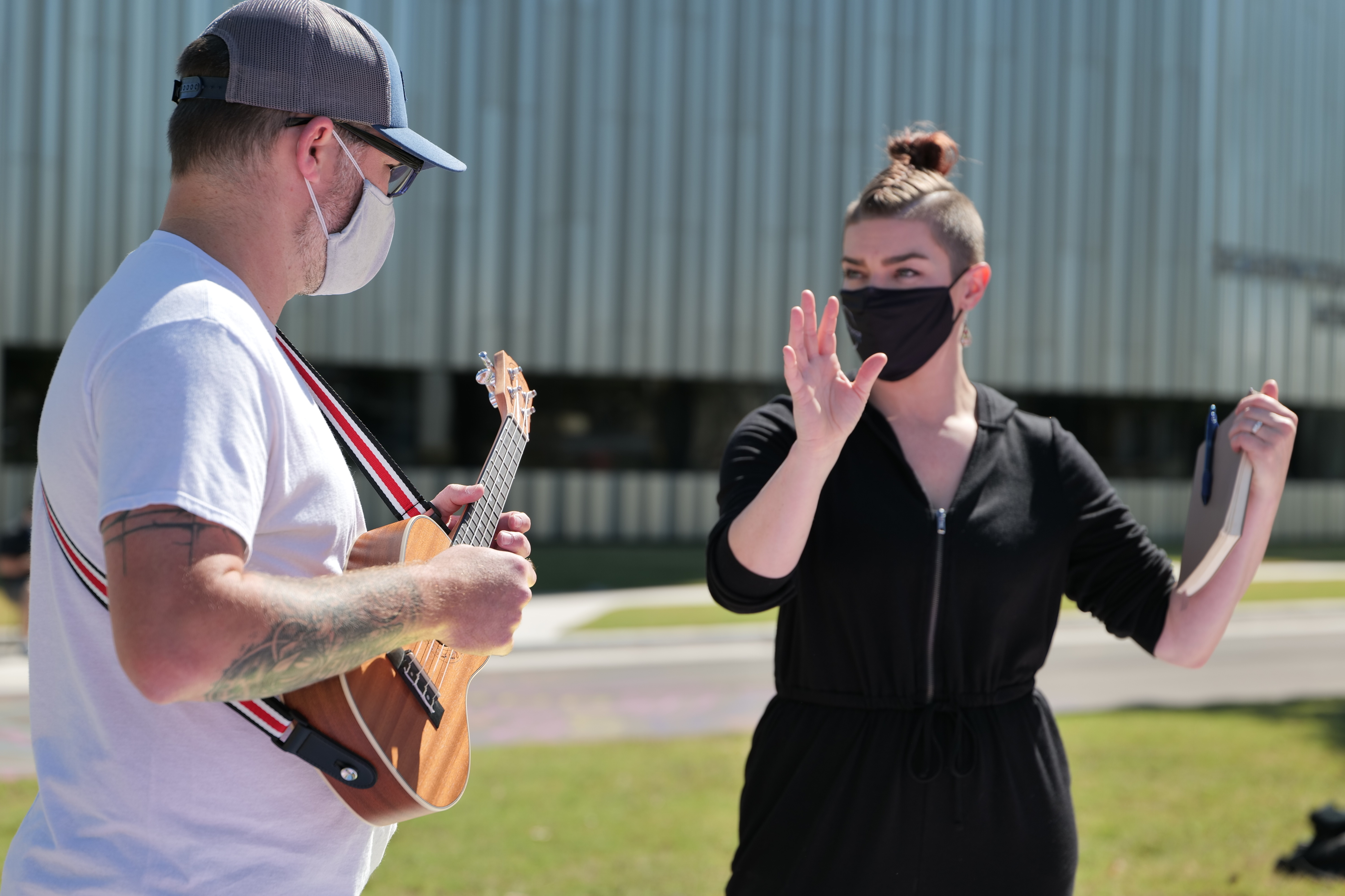 A masked figure holding an acoustic guitar takes direction from a masked figure dressed in black outdoors