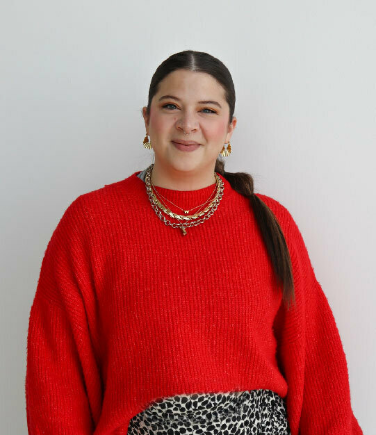 A white woman with long brown hair tied back, wearing chunky gold jewelry and a red sweater tucked into a leopard skirt, stands in front of a white wall smiling