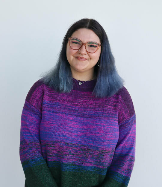 A white woman with black-to-blue short hair, dressed in thick red glasses, with a septum nose ring and striped purple sweater, stands smiling in front of a white wall