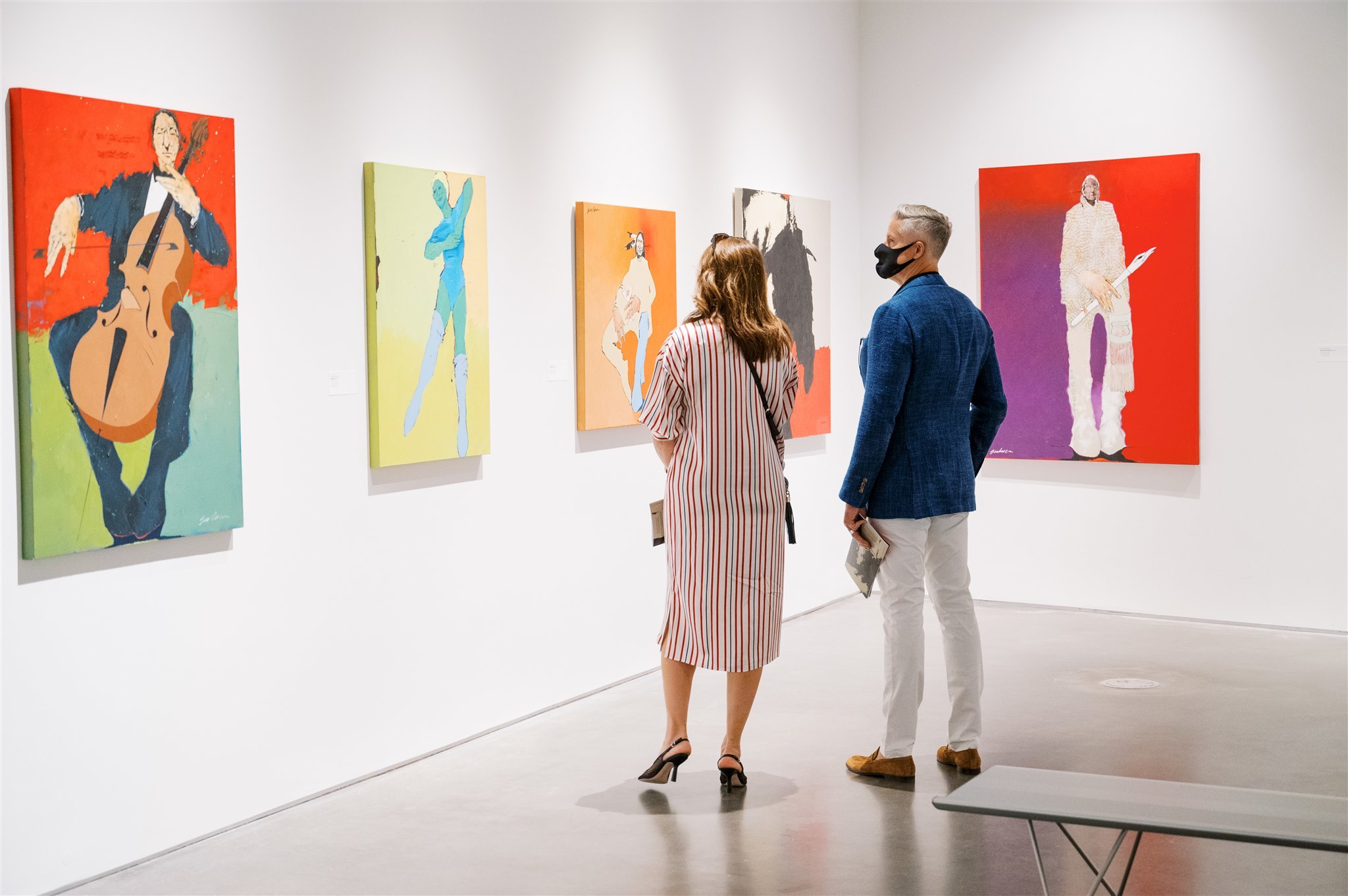 Two people look at paintings hung on white walls in a gallery.