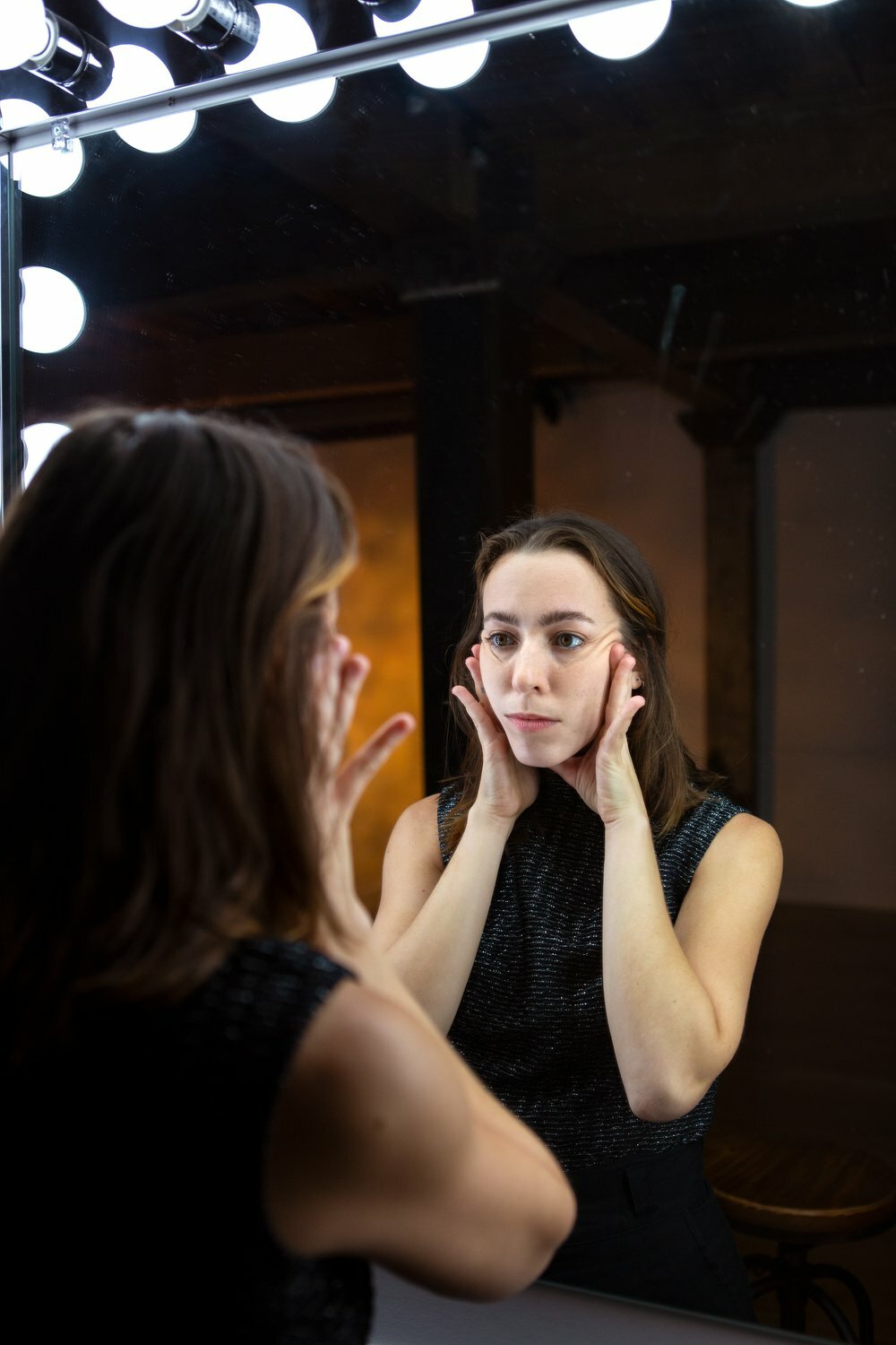 A woman with brown hair holds her face as she examines her reflection in a mirror