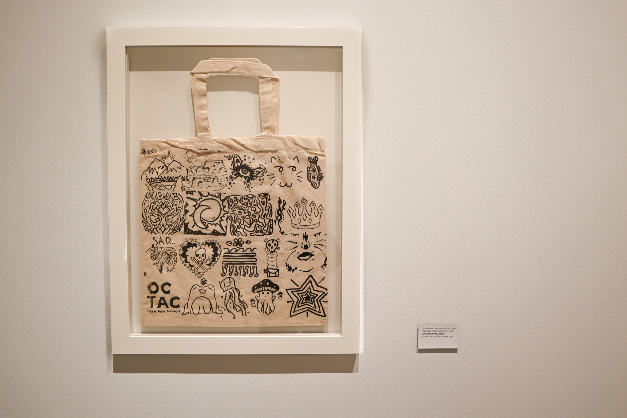 A canvas tote hangs framed on a gallery wall. The tote is decorated in black drawings and doodles.