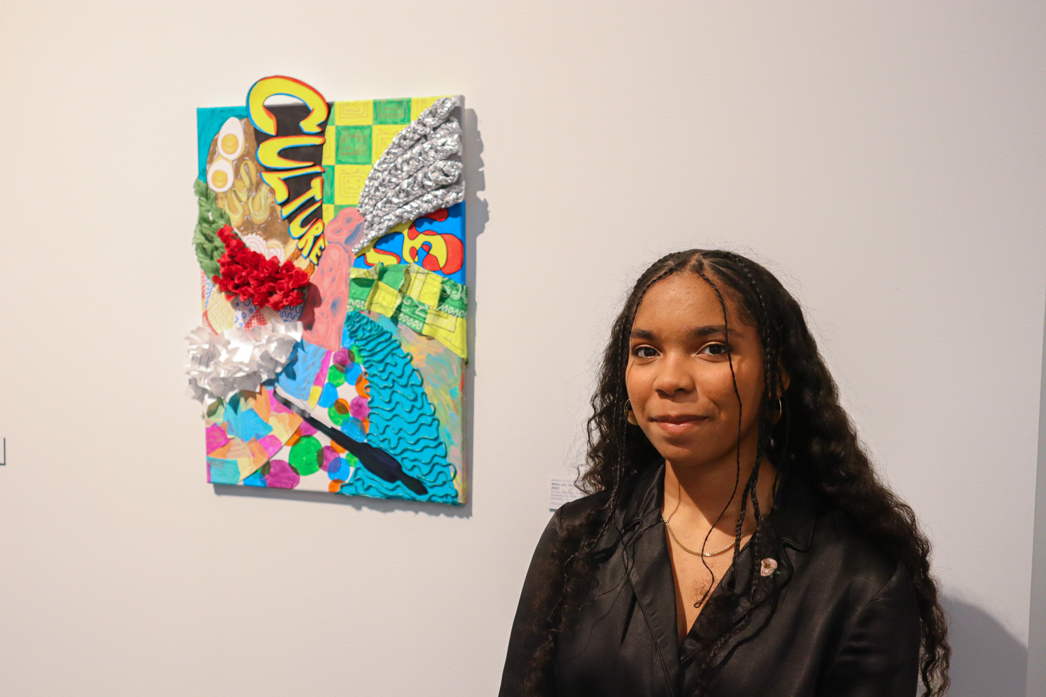 A young person with long dark hair and a black shirt stands in front of a colorful piece of multimedia art hanging on a white gallery wall