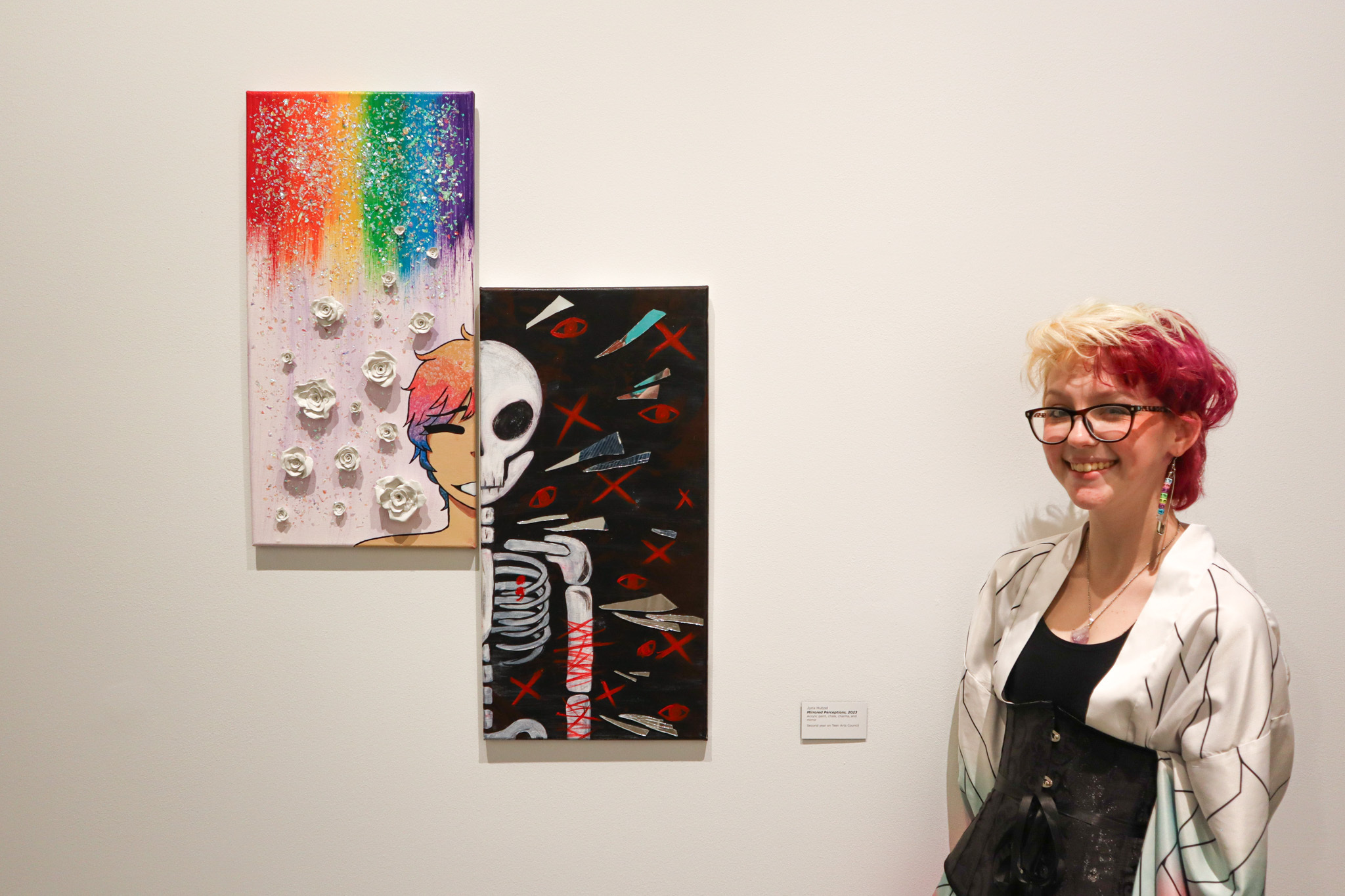 A young person with platinum and bright pink short hair smiles in front of two canvases portraying a rainbow and dark/skeleton side of the same face.