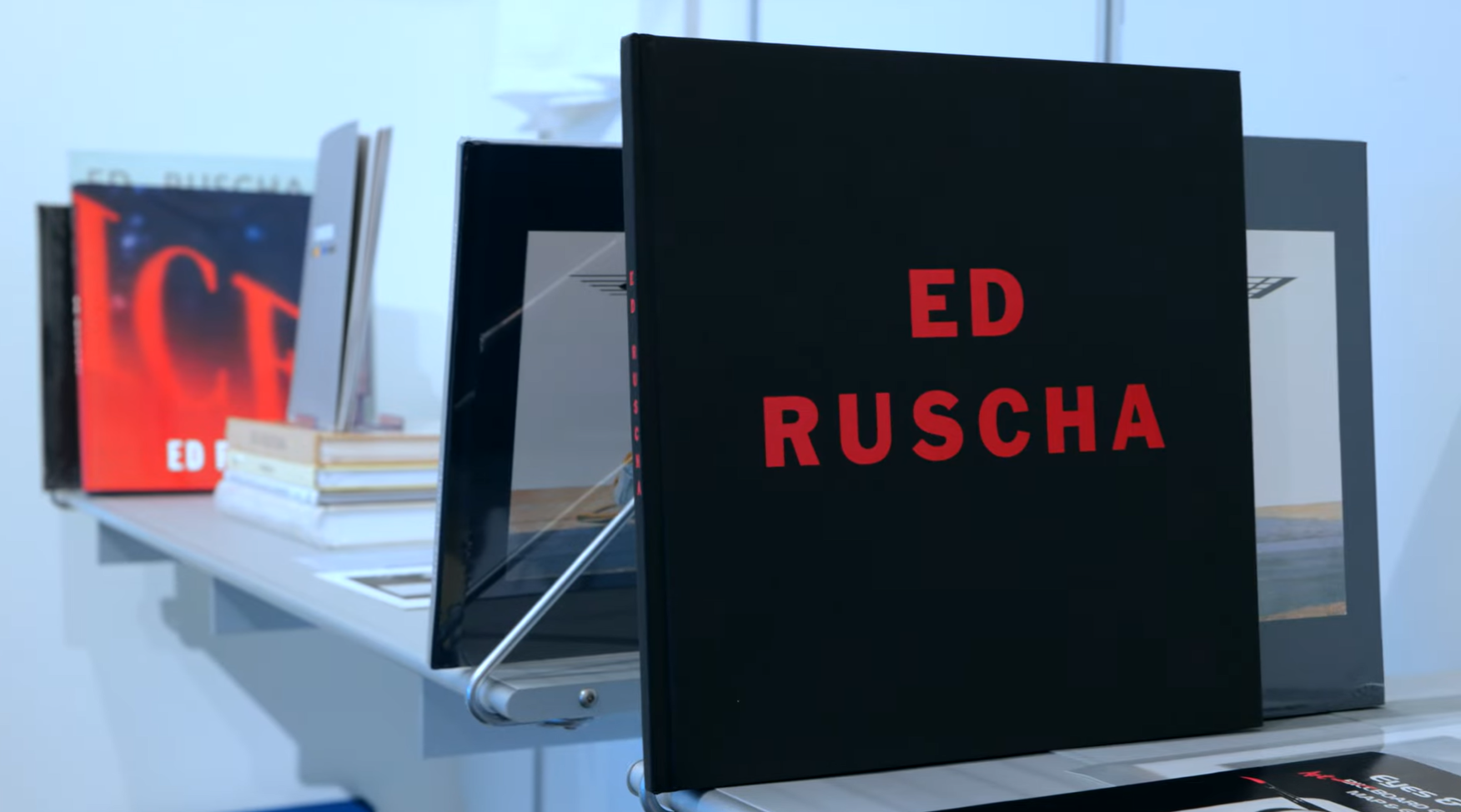 A black book with the words "ED RUSCHA" in red on the cover sits on a shelf with other art books