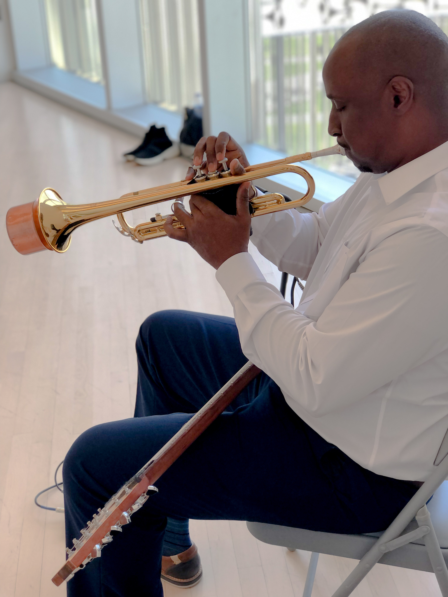 A man in a white dress shirt and blue slacks sits in a chair, showing the camera his profile, by a bright window and is playing the trumpet. We can see a guitar sitting in his lap.