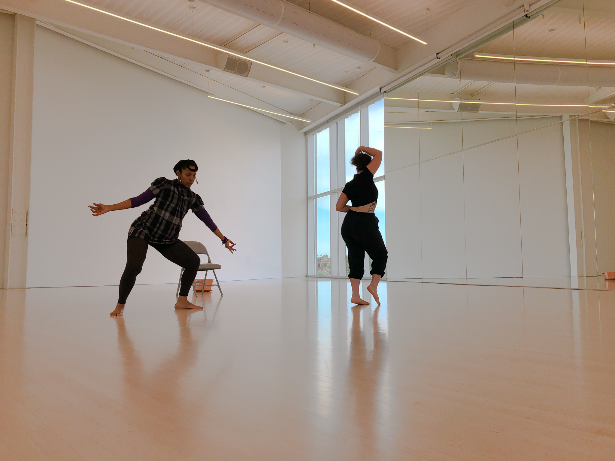 Two dancers are in are in the middle of the rehearsal space, with mirrors and windows on either side. One dancer has her legs spread wide and bent, arms widespread as well. The other is facing away, arms above her head, mid-movement.