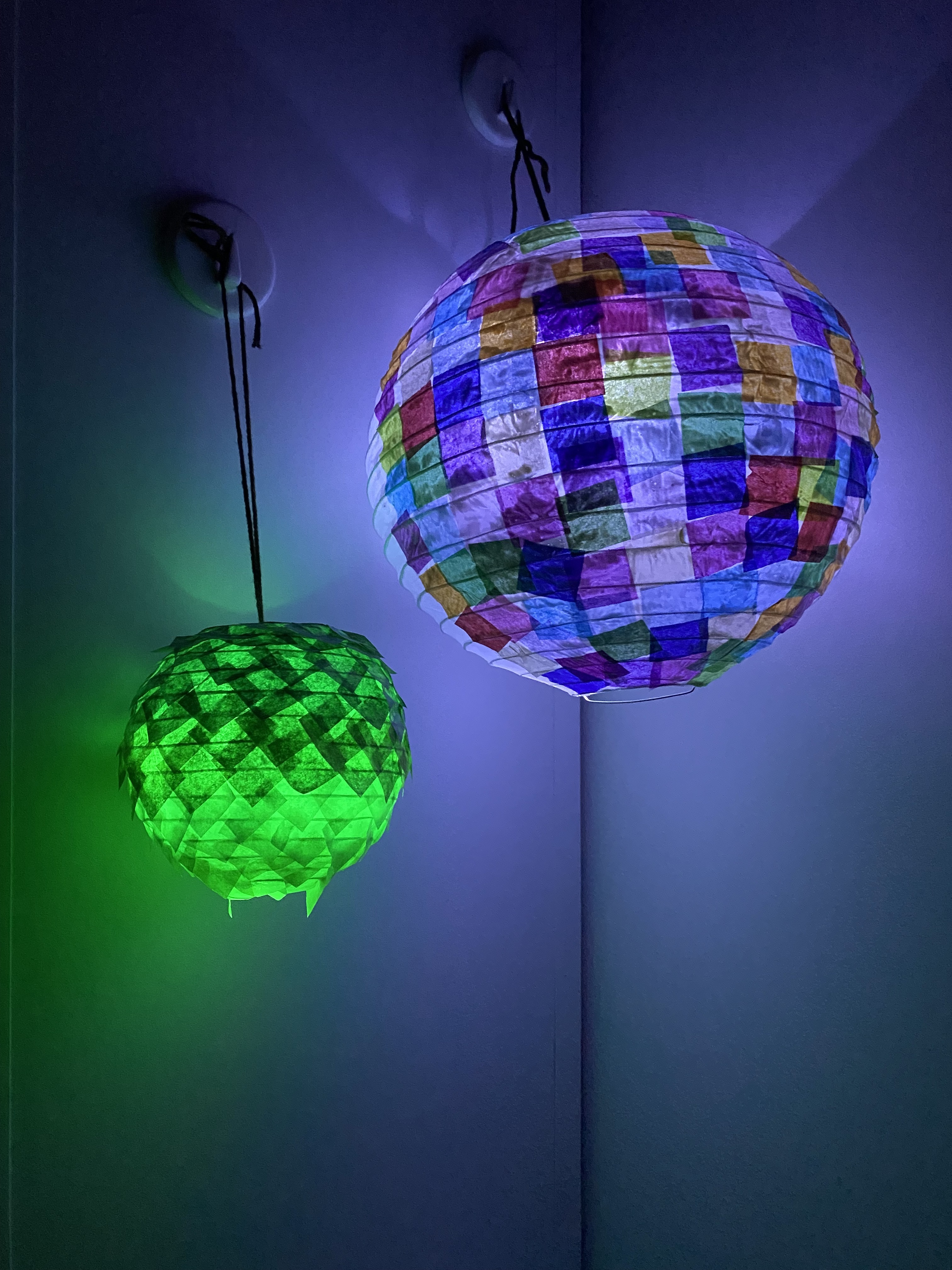 Two paper lanterns -- one green, one mosaic -- hang lit in a darkened room