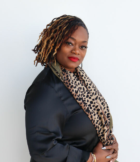 A Black woman with dark-to-light brown locs, a red lip, leopard scarf and black silk shirt smiles softly at the camera
