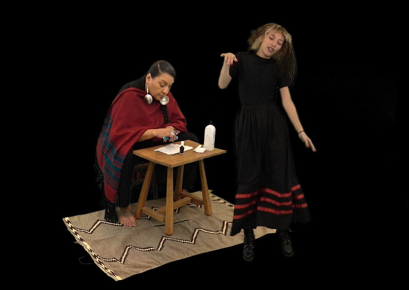 An image with a black background shows one person sitting at a small table and rug drawing. They are dressed in a red blanket with large earrings. A person in a black dress with red stripes at the bottom is dancing in front of the person sitting down.